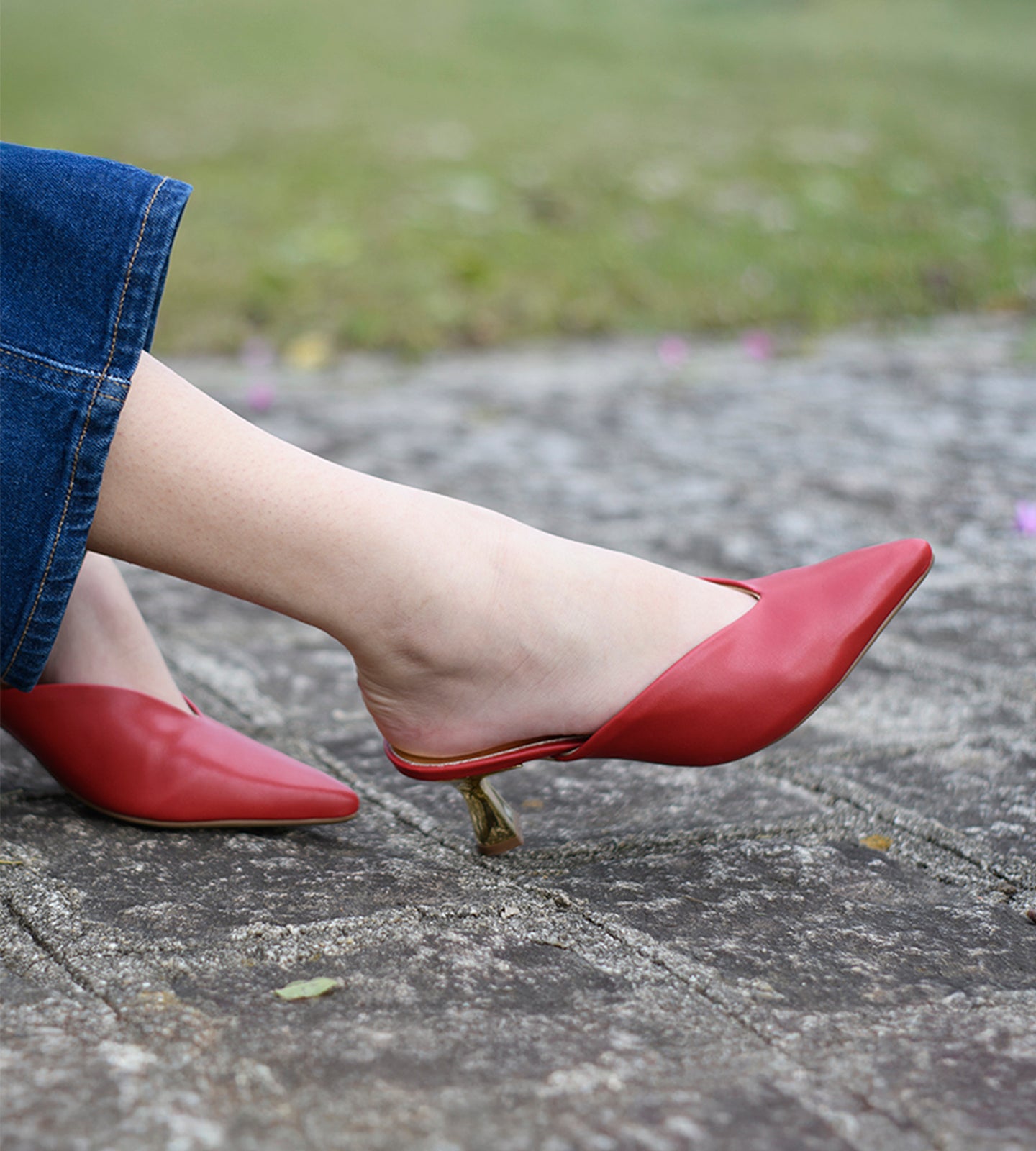 Pessoa usando mule vermelho em napa com bico fino e salto dourado esculpido de 7cm, fotografado em ambiente externo sobre piso de pedra.
