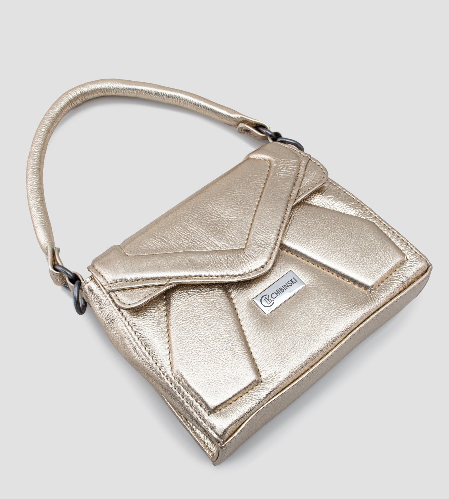 Imagem de uma bolsa feminina pequena da marca Chibinski, confeccionada em couro legítimo dourado. O design moderno apresenta linhas geométricas marcantes, acabamento metálico sofisticado e fecho frontal com a logo da marca em destaque. A bolsa possui alça de mão fixa, ideal para complementar looks elegantes em ocasiões especiais, como festas e casamentos.