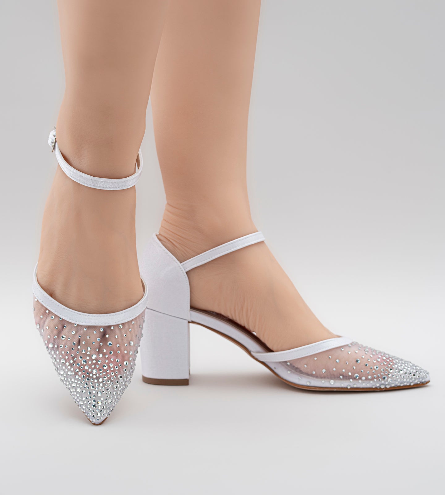 Imagem de um scarpin de noiva branco com salto bloco médio, bico fino e tira com fivela no tornozelo. A parte frontal do sapato é feita em tela delicada com aplicações de pedrarias, criando um efeito brilhante e sofisticado. O calçado combina elegância e conforto, ideal para ocasiões especiais como casamentos e festas.