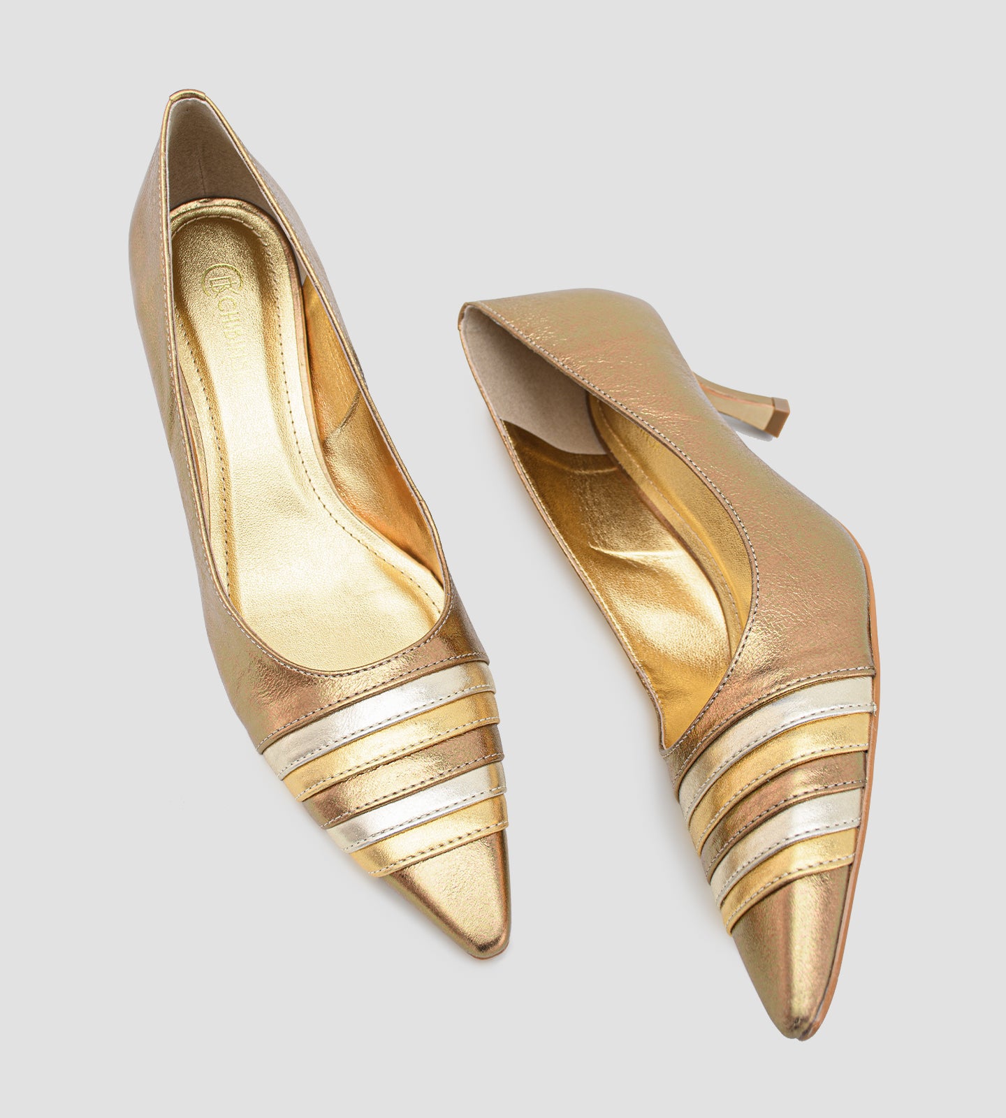 scarpin feminino de couro na cor bronze metalizado. O sapato possui bico fino com um detalhe frontal de tiras metálicas sobrepostas em tons alternados de dourado claro e bronze. O salto é médio, de 7cm, com um design fino que se alarga levemente na base. Fundo liso cinza claro.
