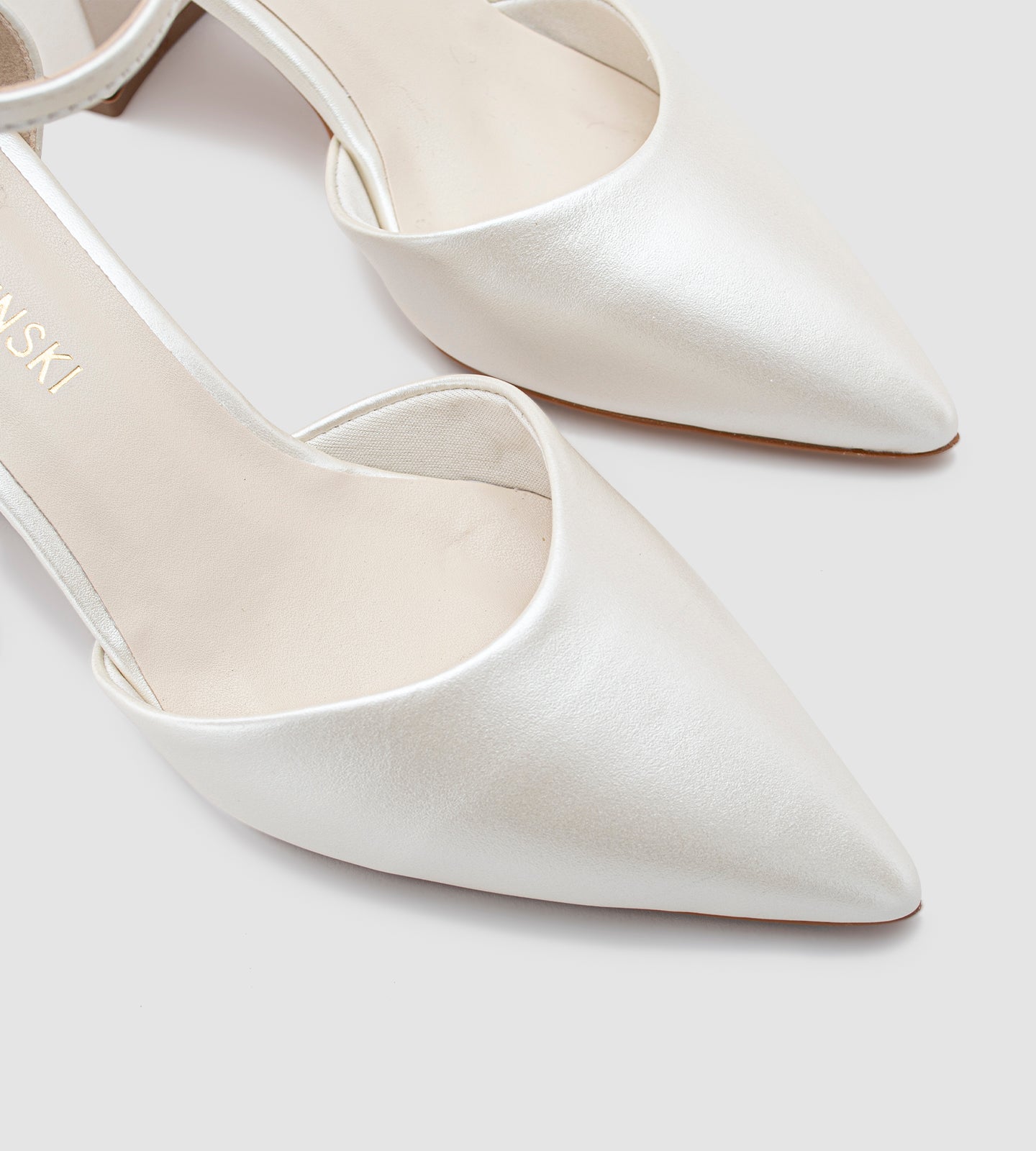 Imagem de um scarpin de noiva na cor off-white com acabamento em couro perolado. O sapato possui bico fino, salto bloco médio e uma tira com fivela dourada ajustável no tornozelo. O design é elegante e minimalista, ideal para casamentos e eventos especiais.