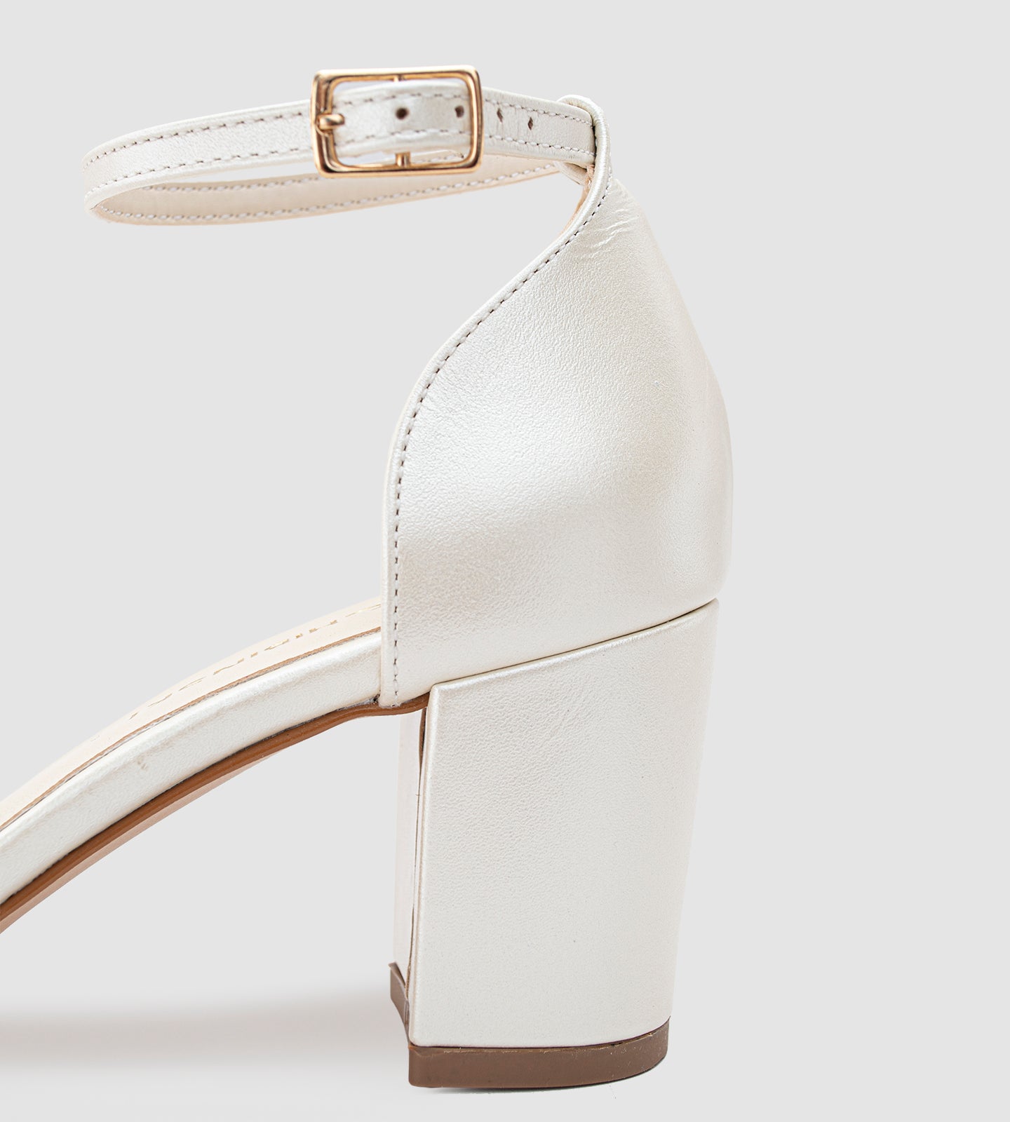 Imagem de um scarpin de noiva na cor off-white com acabamento em couro perolado. O sapato possui bico fino, salto bloco médio e uma tira com fivela dourada ajustável no tornozelo. O design é elegante e minimalista, ideal para casamentos e eventos especiais.