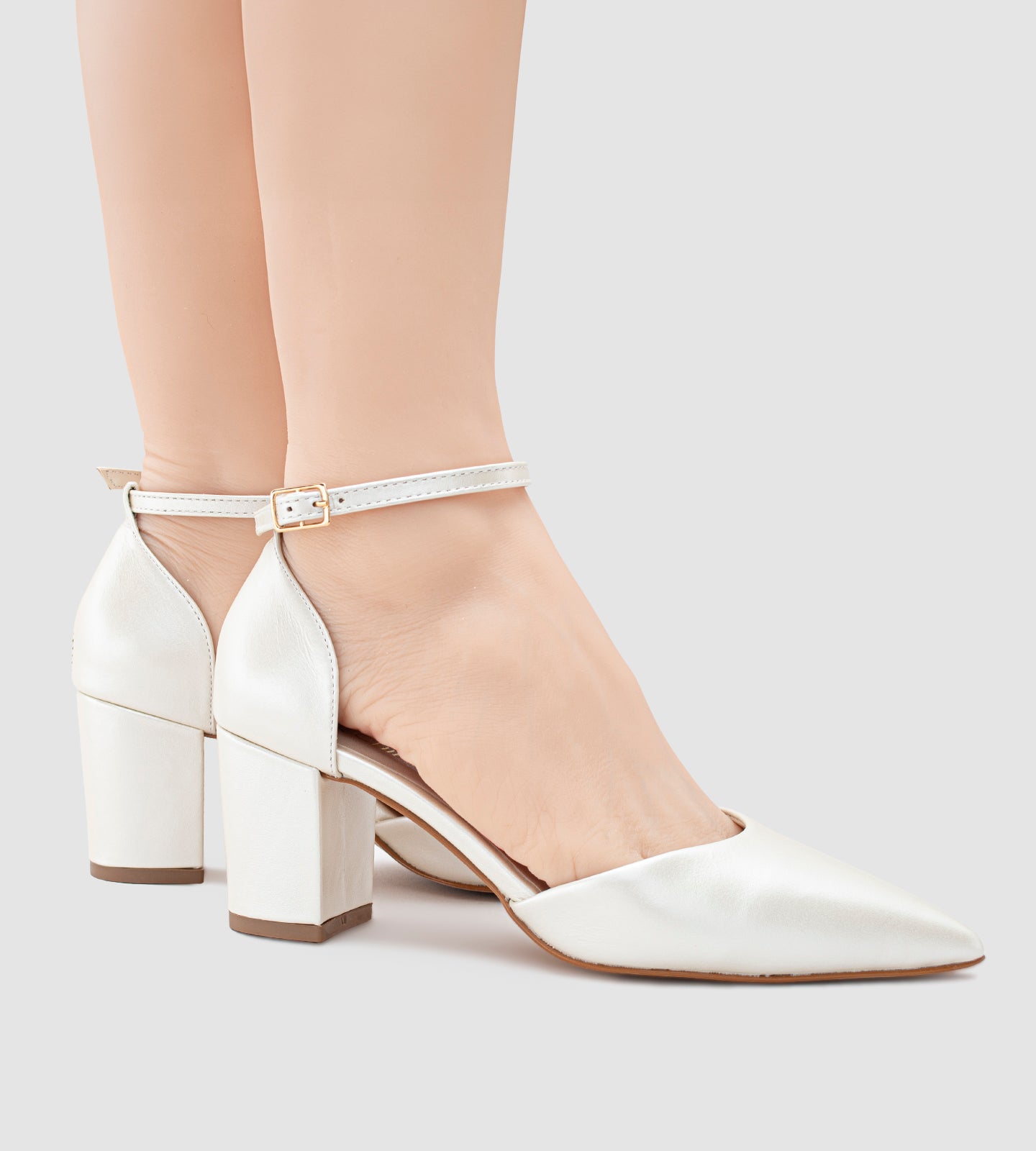 Imagem de um scarpin de noiva na cor off-white com acabamento em couro perolado. O sapato possui bico fino, salto bloco médio e uma tira com fivela dourada ajustável no tornozelo. O design é elegante e minimalista, ideal para casamentos e eventos especiais.