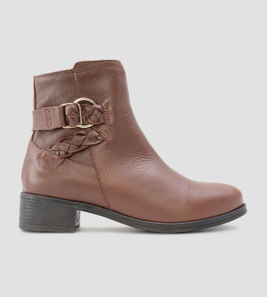 Bota feminina de cano médio em couro marrom com salto bloco baixo. O modelo apresenta detalhe decorativo de tira trançada e fivela dourada no tornozelo, fechamento por zíper lateral e solado emborrachado preto.