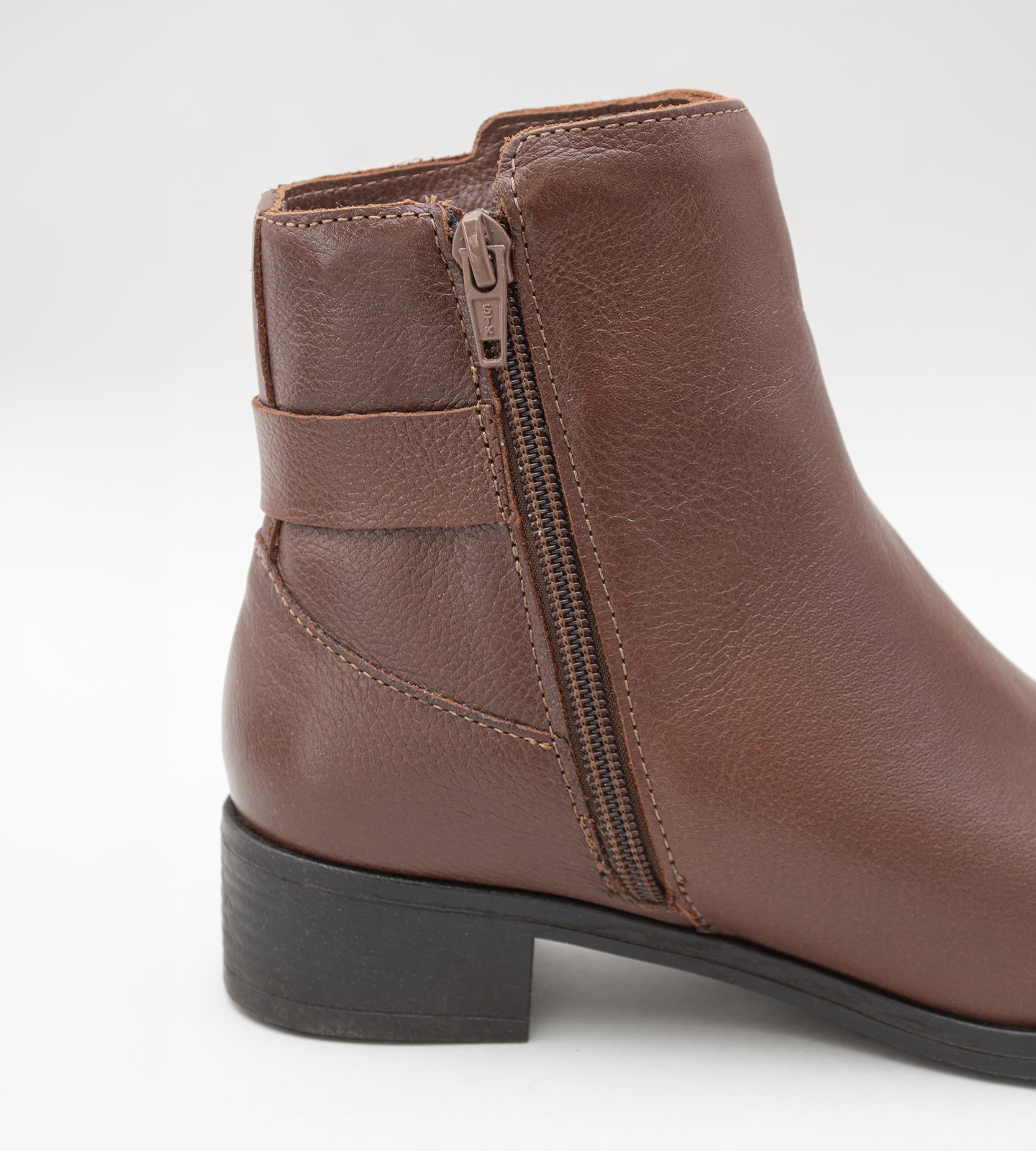 Bota feminina de cano médio em couro marrom com salto bloco baixo. O modelo apresenta detalhe decorativo de tira trançada e fivela dourada no tornozelo, fechamento por zíper lateral e solado emborrachado preto.