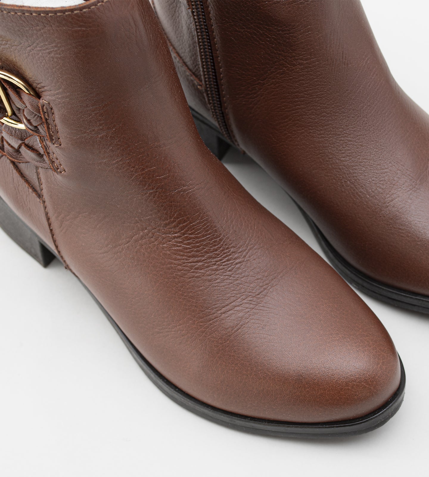 Bota feminina de cano médio em couro marrom com salto bloco baixo. O modelo apresenta detalhe decorativo de tira trançada e fivela dourada no tornozelo, fechamento por zíper lateral e solado emborrachado preto.