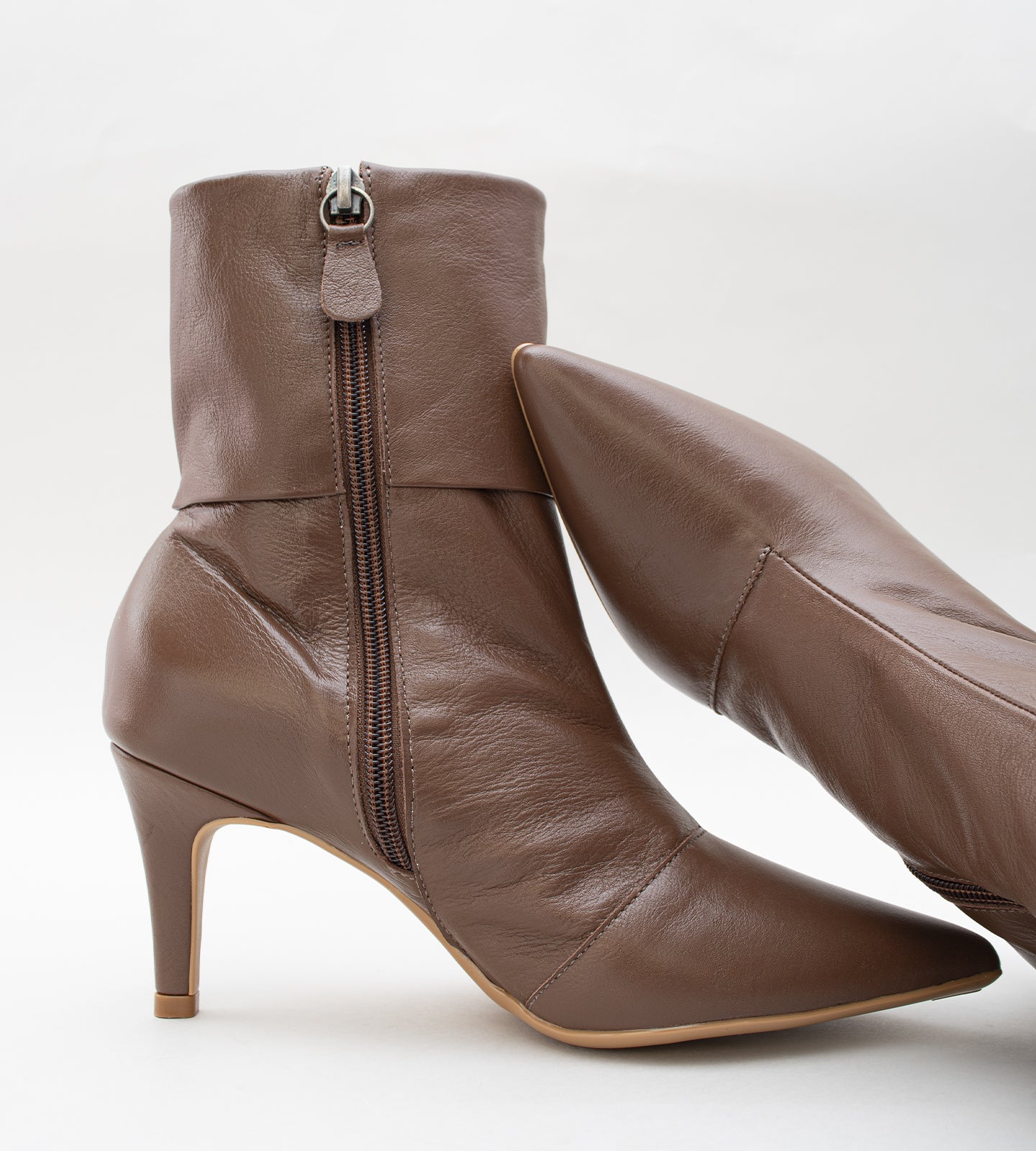 Bota feminina de cano médio em couro marrom. O modelo possui bico fino, salto fino de 8cm e um detalhe de dobra na parte superior do cano.