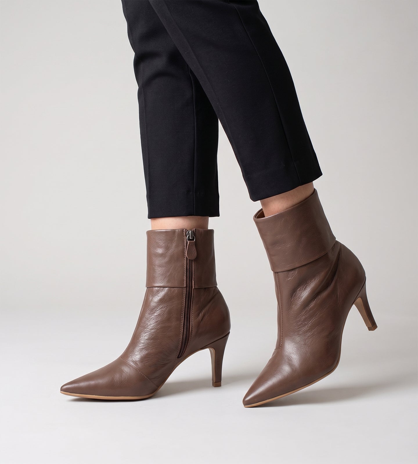 Bota feminina de cano médio em couro marrom. O modelo possui bico fino, salto fino de 8cm e um detalhe de dobra na parte superior do cano.
