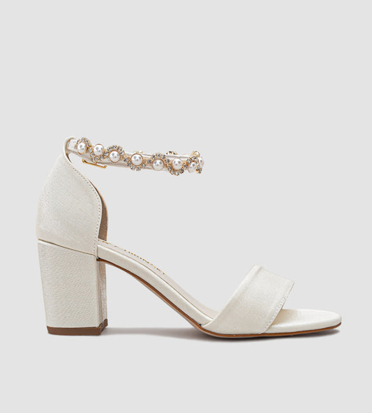 Sandália de noiva off-white em tecido velvet com brilho sutil, salto grosso de 7 cm, tira larga nos dedos e tornozeleira com pérolas e brilhos, unindo conforto, elegância e sofisticação para o grande dia.