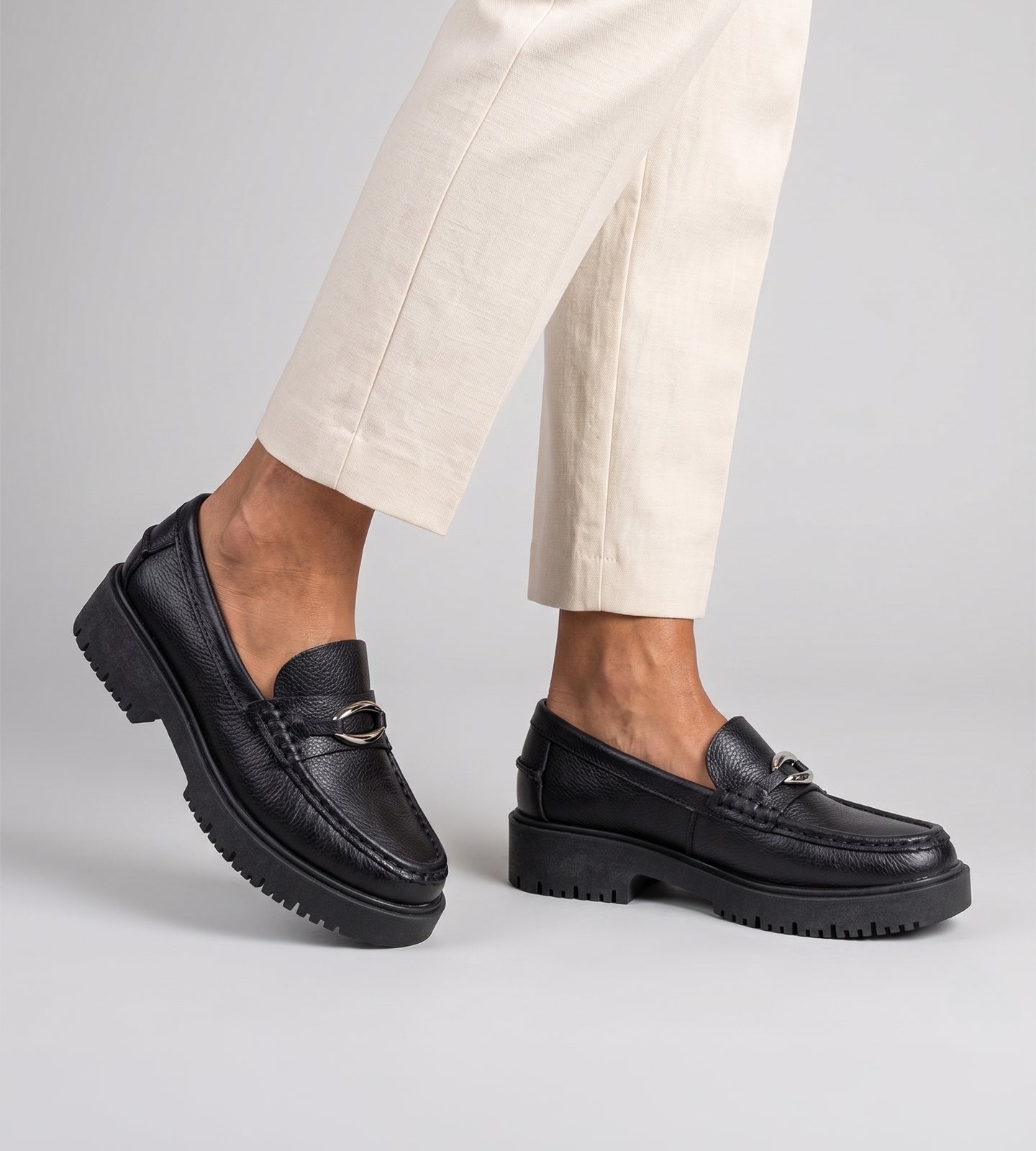 Mocassim feminino em couro preto com solado tratorado e detalhe metálico, usado com calça clara em look moderno.
