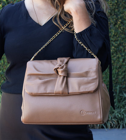 Bolsa feminina em couro marrom com detalhe de nó, corrente dourada e alça ajustável, usada no ombro.