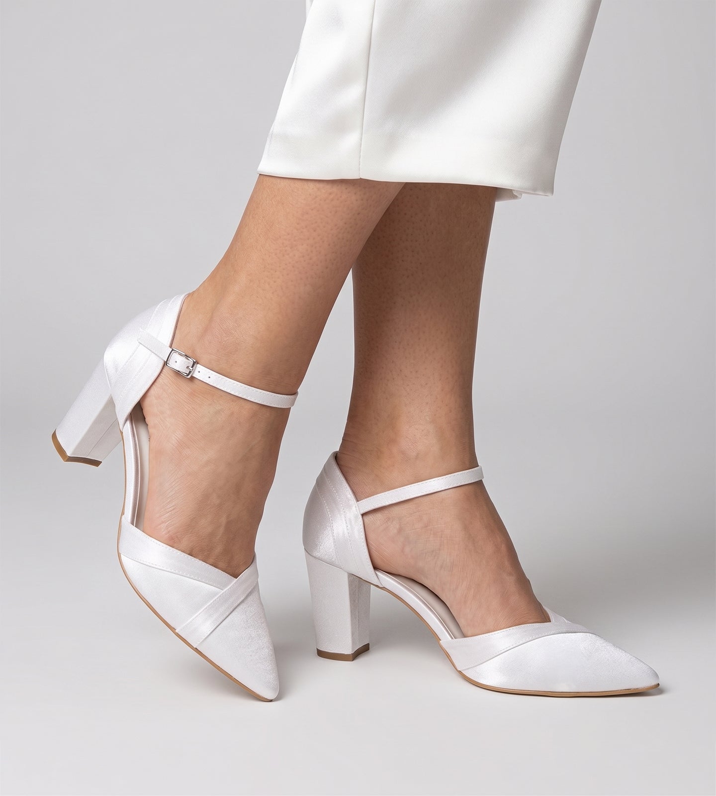 Sapato de noiva scarpin branco em tecido velvet, com bico fino, salto bloco médio de 7 cm e tira no tornozelo com fivela, exibido nos pés com calça clara.