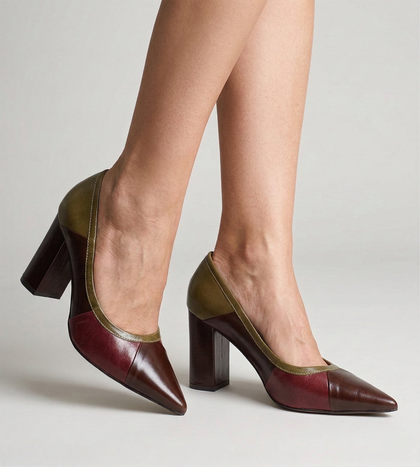 Pernas de uma mulher calçando um scarpin de couro bico fino com salto bloco alto. O sapato possui um design de recortes geométricos em três cores: bico e recortes laterais em marrom escuro, painel em tom de vinho e acabamento do calcanhar e bordas em verde-oliva com costuras claras aparentes. Fundo claro.