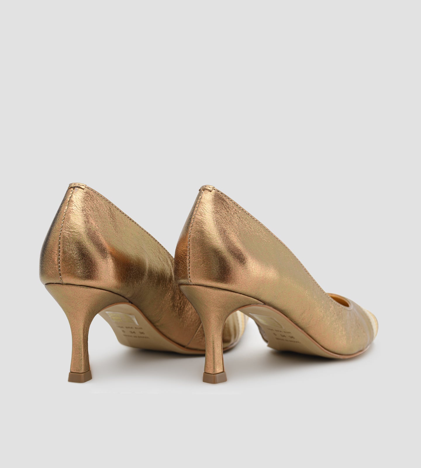 scarpin feminino de couro na cor bronze metalizado. O sapato possui bico fino com um detalhe frontal de tiras metálicas sobrepostas em tons alternados de dourado claro e bronze. O salto é médio, de 7cm, com um design fino que se alarga levemente na base. Fundo liso cinza claro.