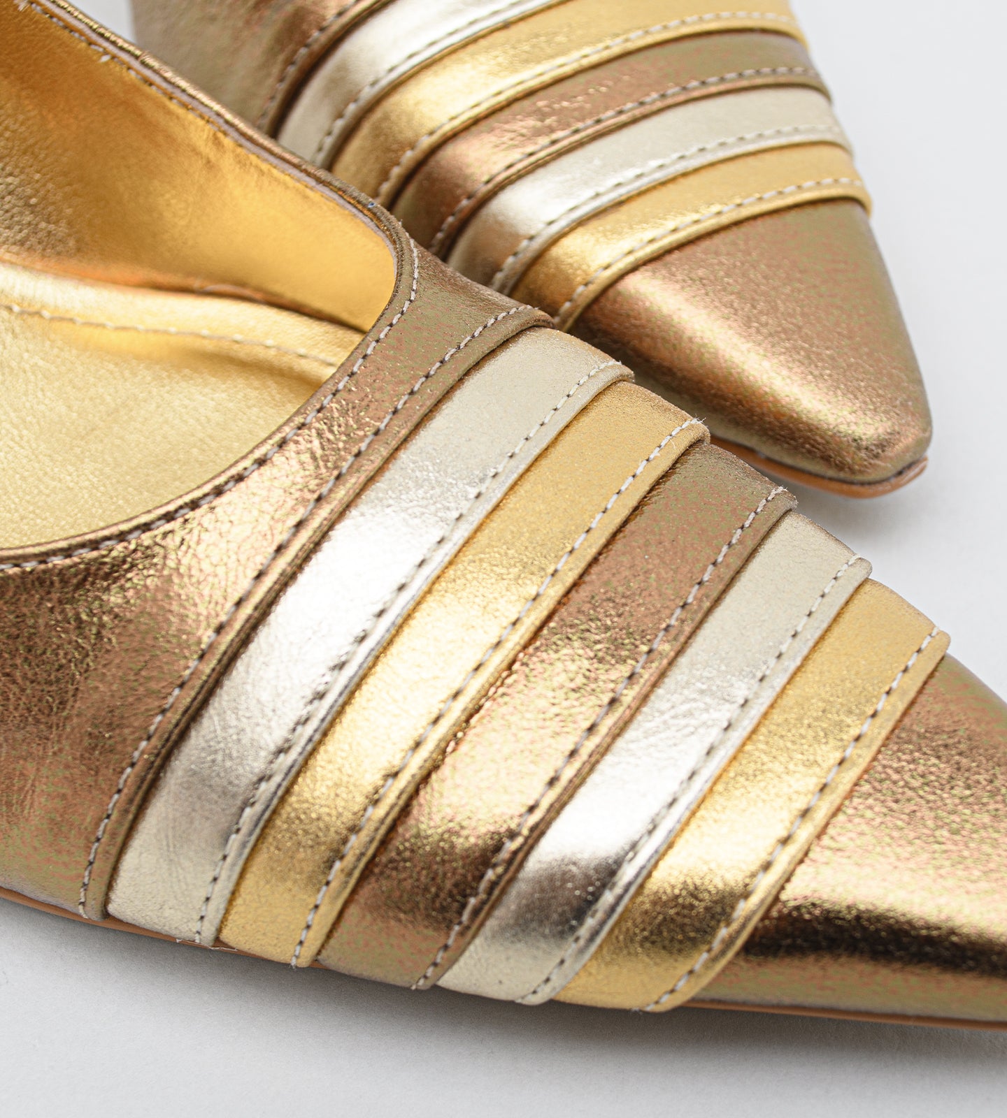 scarpin feminino de couro na cor bronze metalizado. O sapato possui bico fino com um detalhe frontal de tiras metálicas sobrepostas em tons alternados de dourado claro e bronze. O salto é médio, de 7cm, com um design fino que se alarga levemente na base. Fundo liso cinza claro.