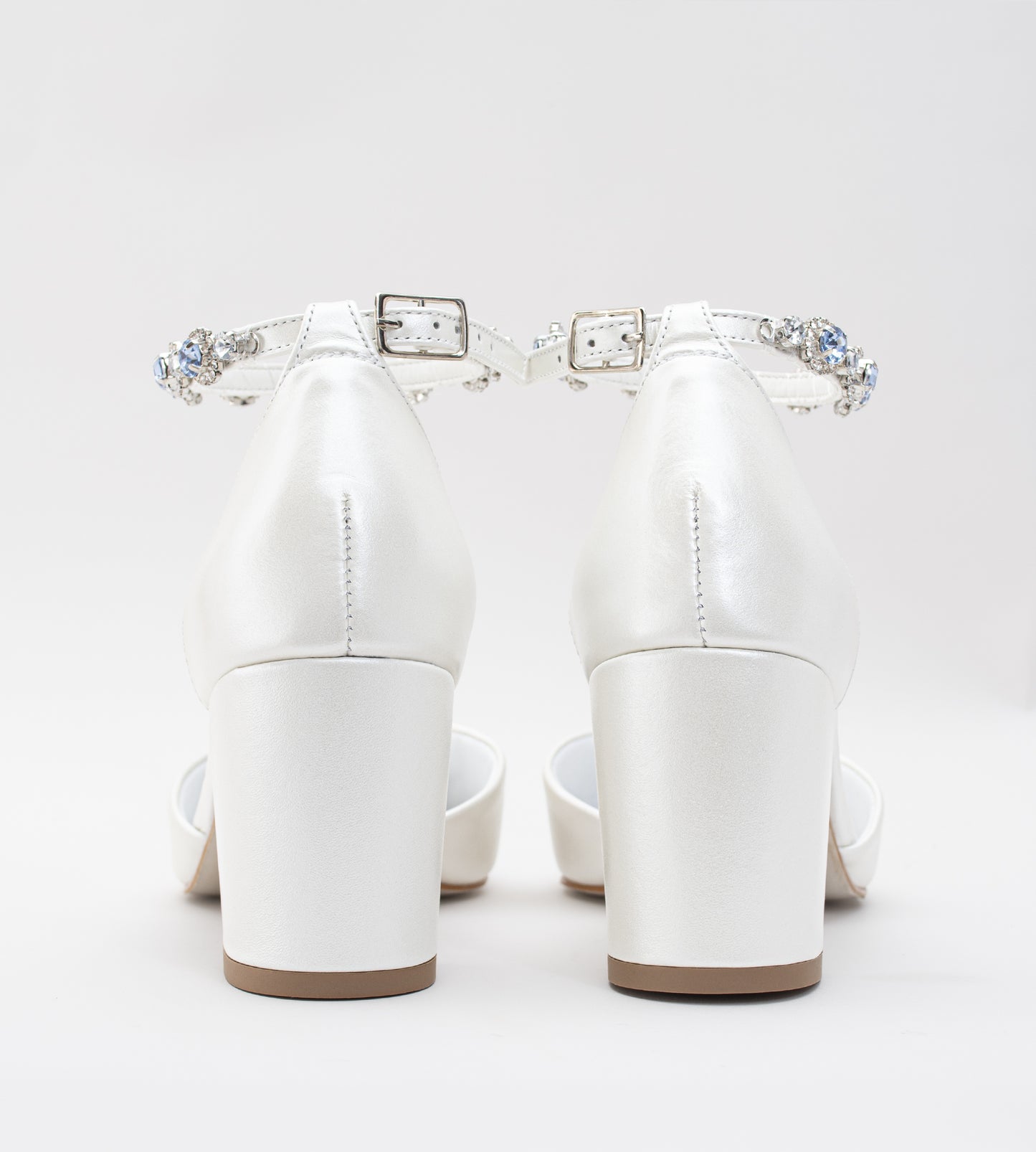 Sapato de noiva em couro branco perolado com bico fino, salto bloco médio e pulseira no tornozelo adornada com cristais azul-claro, ideal para casamento, fotografado em fundo cinza claro.