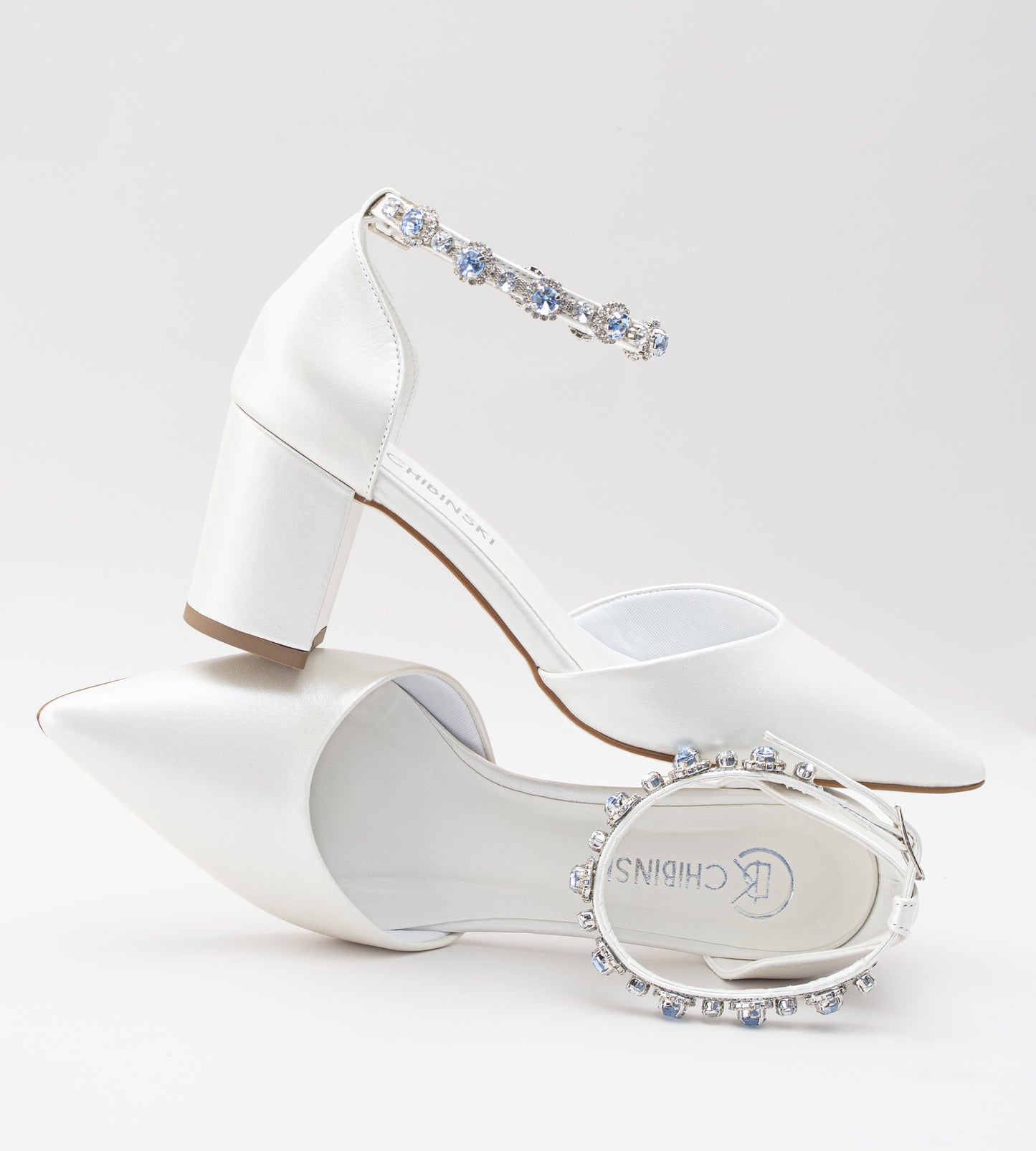 Sapato de noiva em couro branco perolado com bico fino, salto bloco médio e pulseira no tornozelo adornada com cristais azul-claro, ideal para casamento, fotografado em fundo cinza claro.