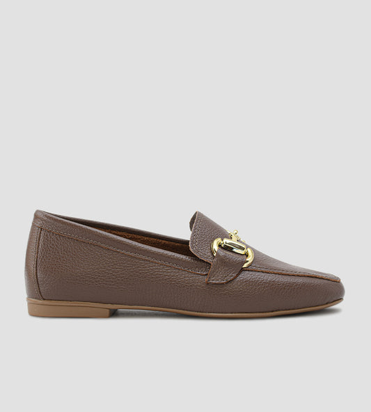 Mocassim Loafer feminino em couro legítimo marrom, com bico levemente quadrado/fino, solado baixo e detalhe de bridão (fivela) metálico dourado no peito do pé. O sapato possui costura aparente e acabamento em couro texturizado.