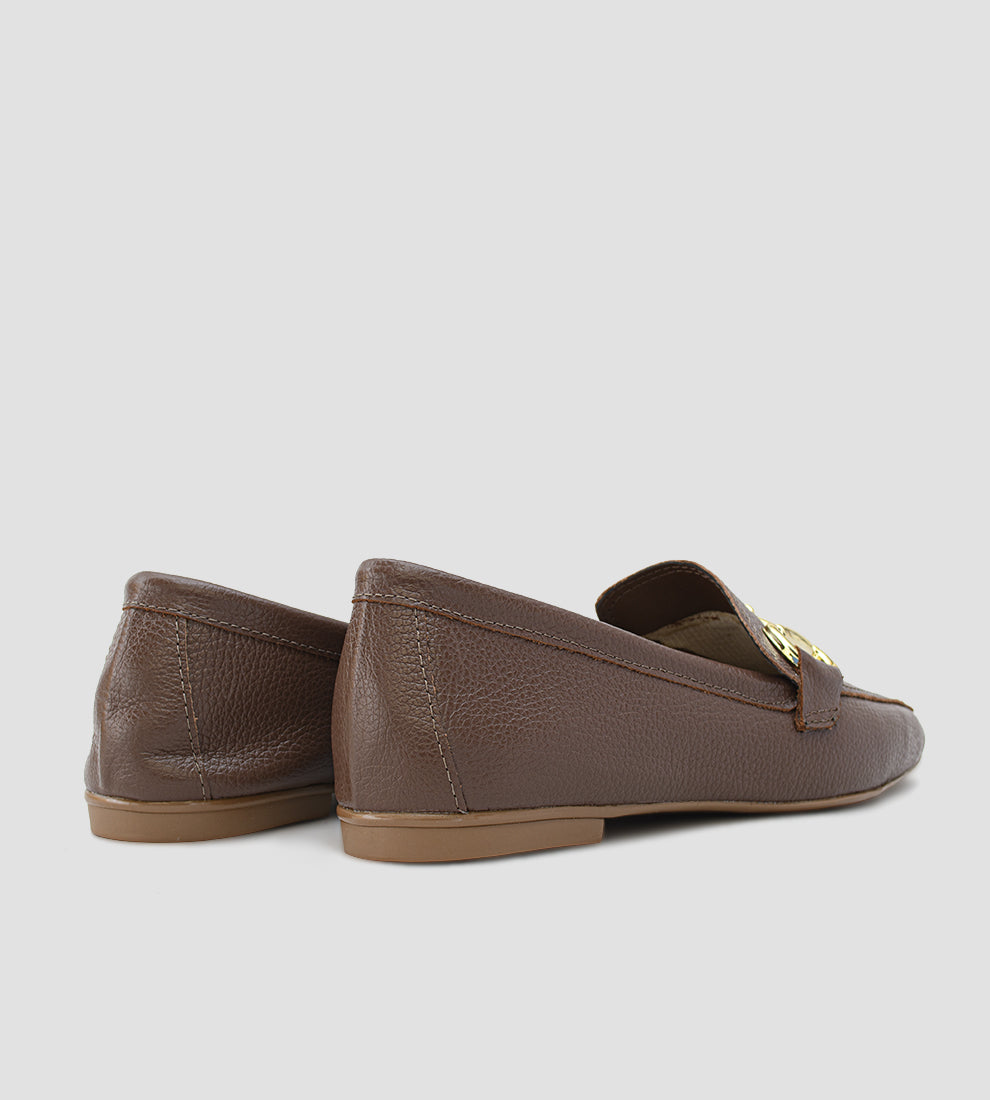 Mocassim Loafer feminino em couro legítimo marrom, com bico levemente quadrado/fino, solado baixo e detalhe de bridão (fivela) metálico dourado no peito do pé. O sapato possui costura aparente e acabamento em couro texturizado.