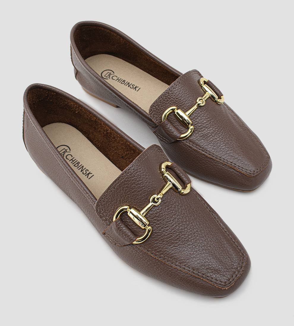 Mocassim Loafer feminino em couro legítimo marrom, com bico levemente quadrado/fino, solado baixo e detalhe de bridão (fivela) metálico dourado no peito do pé. O sapato possui costura aparente e acabamento em couro texturizado.