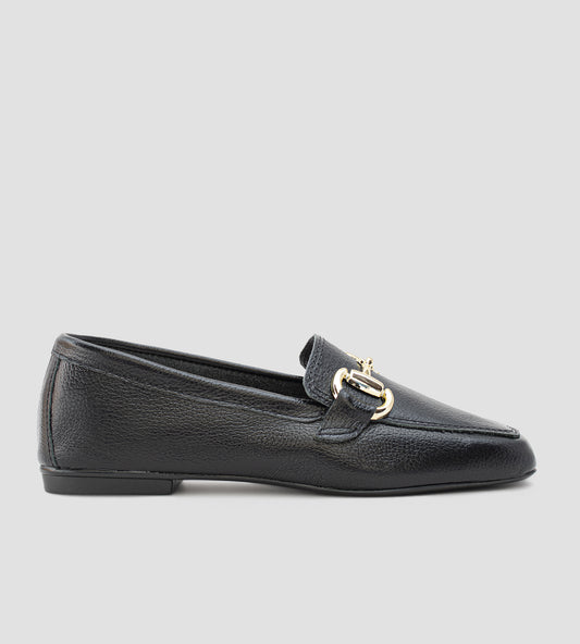 Mocassim feminino preto em couro legítimo com detalhe metálico dourado frontal e solado baixo, usado com calça clara em composição elegante e sofisticada.