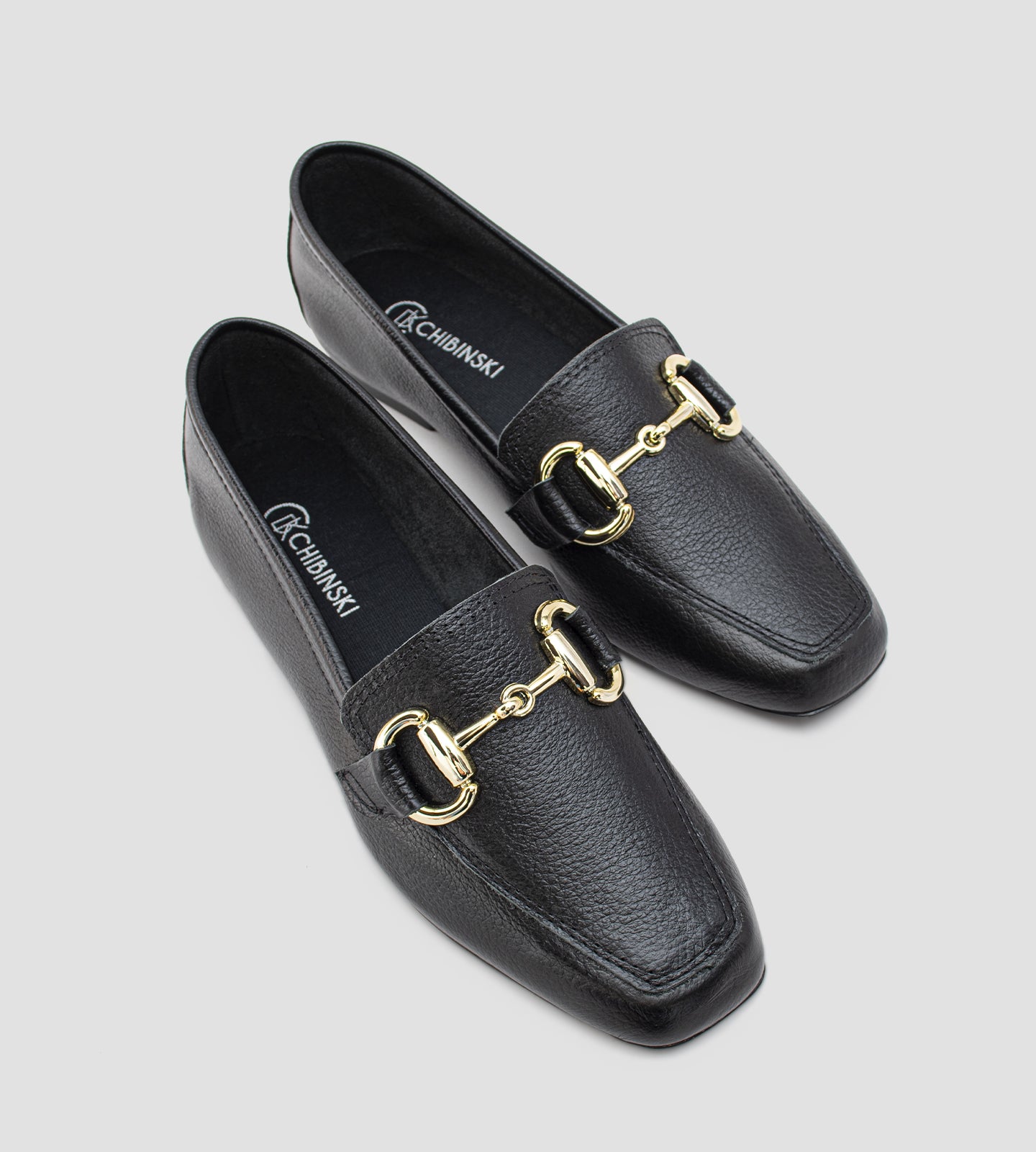 Mocassim feminino preto em couro legítimo com detalhe metálico dourado frontal e solado baixo, usado com calça clara em composição elegante e sofisticada.