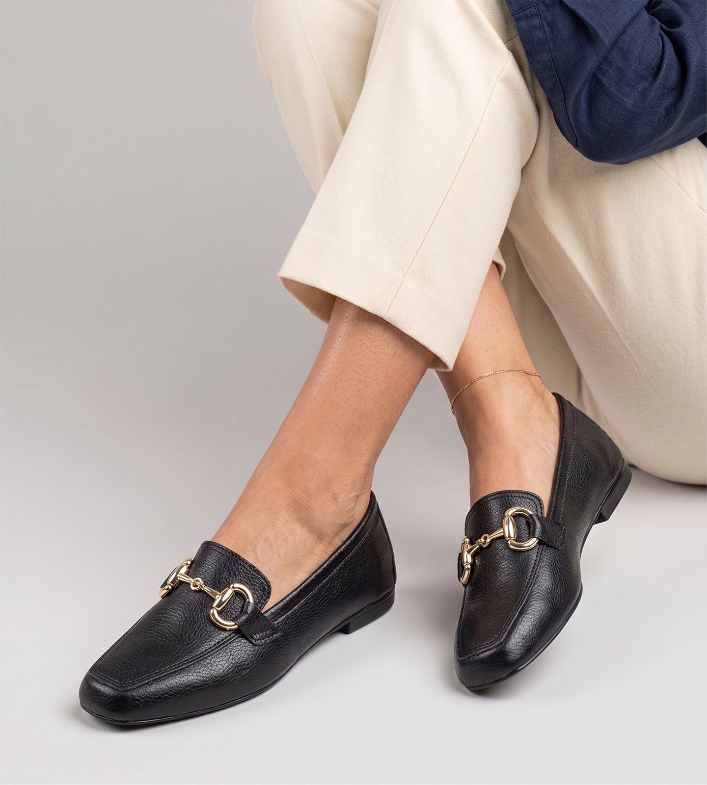 Mocassim feminino preto em couro legítimo com detalhe metálico dourado frontal e solado baixo, usado com calça clara em composição elegante e sofisticada.