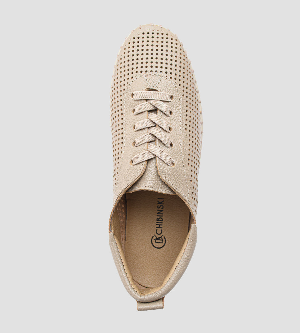 Tênis Feminino Casual na cor nude/bege claro, com o cabedal totalmente perfurado e cadarços finos. O tênis possui solado flexível estilo driving shoe, emborrachado e costurado, com um dos pés dobrado para demonstrar a alta flexibilidade.